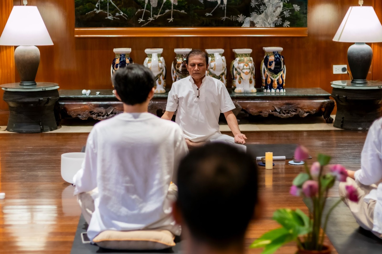 A back-view of practitioners meditating on mats, facing a wellness master in white attire who is leading a meditation session in a warm, elegantly decorated wooden room.