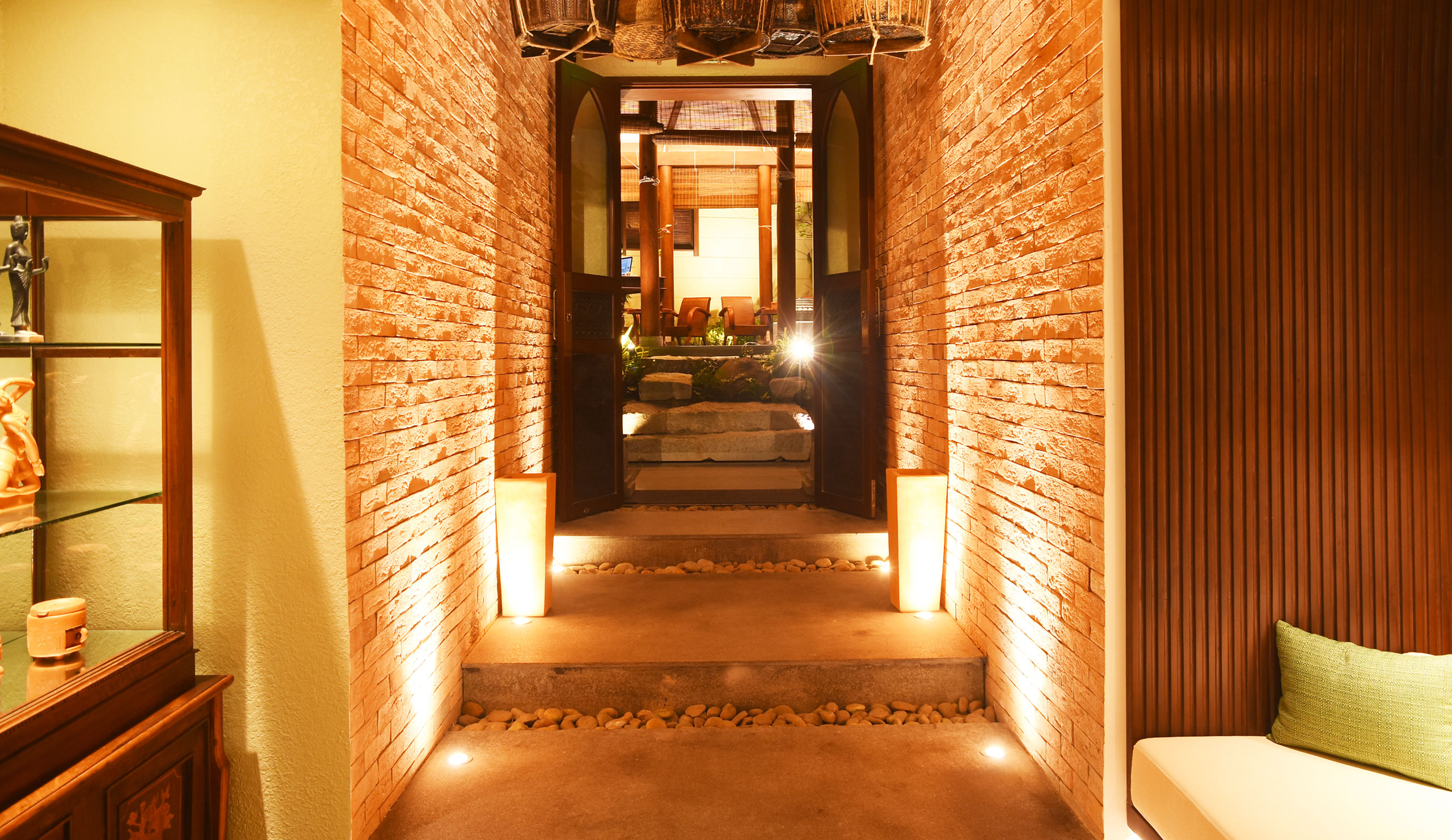 Một hành lang lối vào được chiếu sáng đẹp mắt tại V-Senses Wellness & Spa với những bức tường gạch mộc, ánh sáng sàn dịu nhẹ dọc theo các bậc thang và những cánh cửa gỗ mở dẫn vào một sân vườn chăm sóc sức khỏe yên bình.